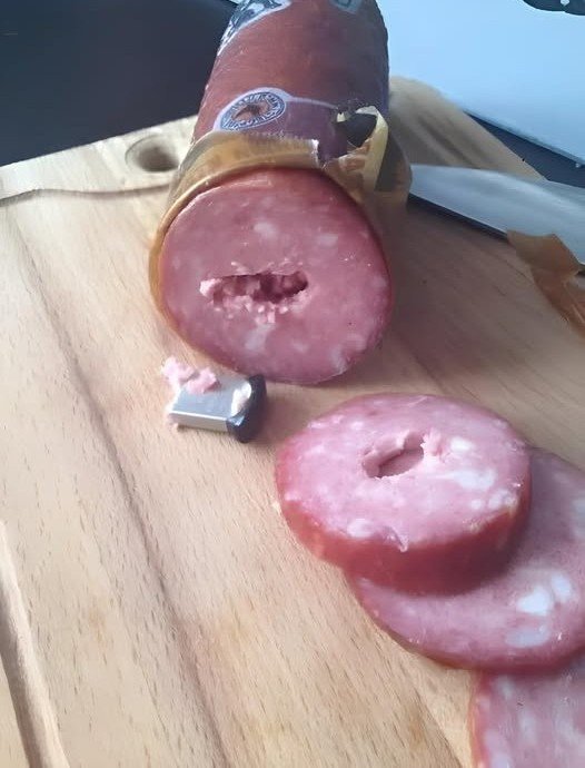 Χθες αγόρασα από το κατάστημα ένα συνηθισμένο σαλάμι∙ τίποτα ιδιαίτερο. Ήθελα απλώς να ετοιμάσω μερικά σάντουιτς. Όταν γύρισα σπίτι, έκοψα μερικές φέτες, τις έφαγα και έβαλα το υπόλοιπο στο ψυγείο. Όλα έμοιαζαν απολύτως φυσιολογικά. Μέχρι που ξαφνικά ανακάλυψα μια μονάδα USB μέσα στο σαλάμι. Αρχικά νόμιζα ότι είχε πέσει εκεί κατά λάθος, όμως η περιέργεια με κυρίευσε και αποφάσισα να δω τι περιείχε. Το επόμενο πρωί, καθώς ετοίμαζα το πρωινό, πήρα ξανά το σαλάμι και άρχισα να το κόβω. Τότε παρατήρησα κάτι παράξενο: η υφή του δεν ήταν ίδια, σαν να υπήρχε κάτι σκληρό στο εσωτερικό. Σκέφτηκα ότι ίσως ήταν απλώς πολύ παγωμένο. Όμως όταν προσπάθησα να κόψω άλλη μια φέτα, το μαχαίρι σταμάτησε απότομα. Έσκυψα να κοιτάξω καλύτερα… και πάγωσα. Κάτι μεταλλικό γυάλιζε μέσα στο λουκάνικο. Αρχικά νόμισα πως ήταν κάποιο κομμάτι μετάλλου, αλλά όταν το κούνησα ελαφρά, μια μονάδα USB ξεπρόβαλε από το ροζ κρέας. Μια απλή κάρτα μνήμης, με λίγα gigabytes χωρητικότητας. Και το χειρότερο; Είχα ήδη φάει μέρος από αυτό το σαλάμι. Πώς ήταν δυνατόν να βρεθεί μια USB μέσα σε ένα βιομηχανικά συσκευασμένο προϊόν — και μάλιστα όχι φθηνό; Παρά τον φόβο μου, η περιέργεια υπερίσχυσε. Άνοιξα τον υπολογιστή, σύνδεσα τη μονάδα USB και κοίταξα τα αρχεία της. Υπήρχε μόνο ένας φάκελος, με τίτλο «ΑΝΟΙΞΕ ΜΕ». Τον άνοιξα. Μέσα υπήρχε μία και μοναδική εικόνα: ένας άντρας που κοιτούσε την κάμερα και χαμογελούσε. Ένιωσα ένα ρίγος να με διαπερνά. Άφησα το ποντίκι και έμεινα να κοιτάζω την οθόνη ακίνητος. Ίσως να ήταν κάποιο κακόγουστο αστείο… αλλά τότε γιατί να κρύψει κάποιος μια μονάδα USB μέσα σε ένα σαλάμι; Και σε ποιον προοριζόταν; Δεν ξέρω αν πρέπει να πάω στην αστυνομία για να ανακαλύψω ποιος κρύβεται πίσω από όλο αυτό ή απλώς να πετάξω το καταραμένο σαλάμι και να προσπαθήσω να το ξεχάσω. Είναι σίγουρα μια παράξενη ιστορία — αλλά με έναν περίεργο τρόπο, και γελοία.